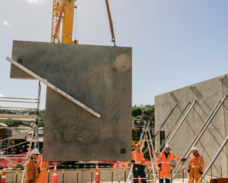 A prefabricated concrete wall is being carefully lowered onto the foundations so it aligns with the steel rods that it must fit onto. There are five construction workers in high vis overalls and hard hats watching and helping to guide it.