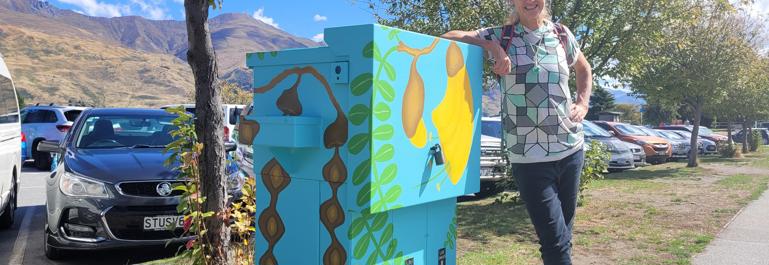 Streetart: Wānaka artist, Chrissy Wickes standing next to a switchbox painted with kowhai at various stages of flowering and seeding