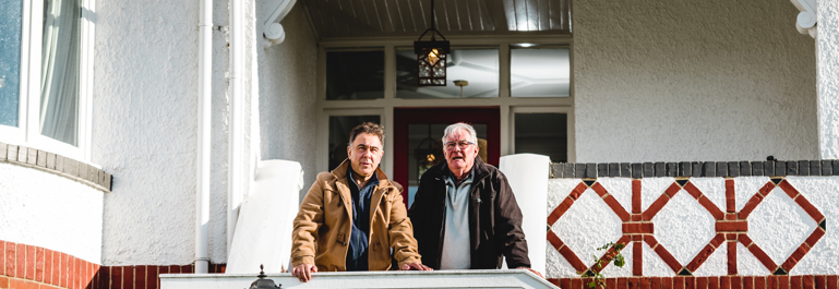 Photo of two men standing on the steps of a brick house leaning on the handrail looking toward the camera.