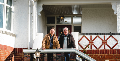Photo of two men standing on the steps of a brick house leaning on the handrail looking toward the camera.