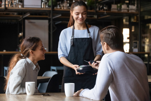 Image shows two people sitting at a table in a cafe with coffee mugs, one of them is paying the aproned waitress who is holding a mobile EFTPOS machine.waitress