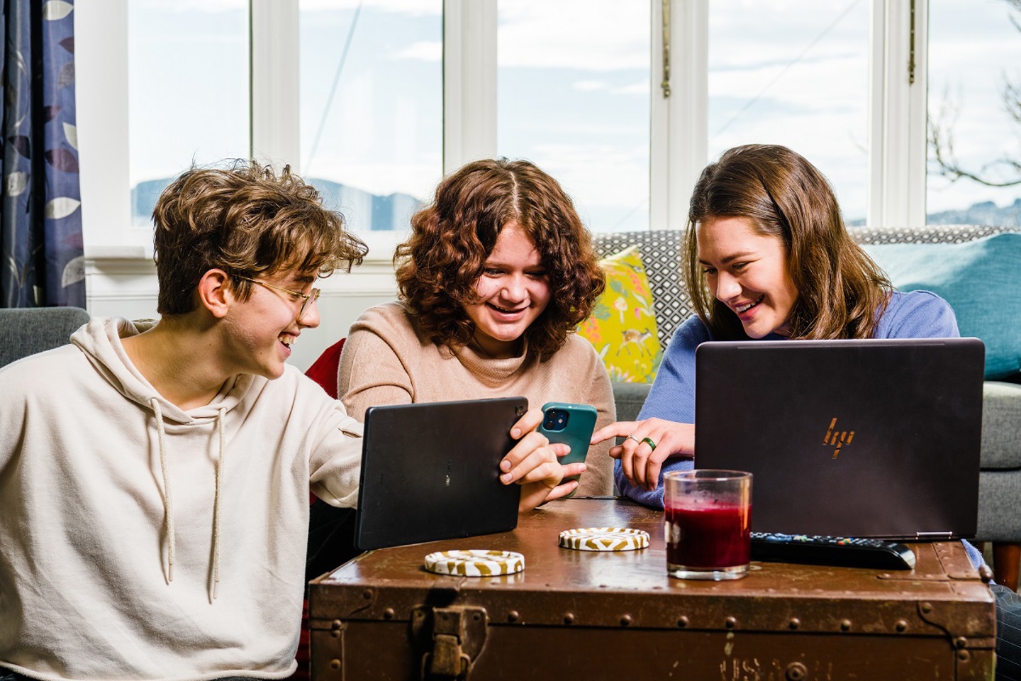 Three young adults sitting around a coffee table using devices including tablets, phones and laptops.