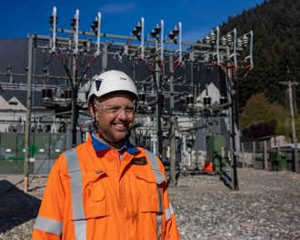 Photo shows a person in PPE gear in the foreground in front of a substation.