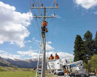 Photo shows two workers in a cherry picker basket with another worker on a ladder with safety harness at the top of a power pole working.