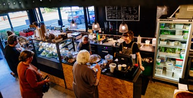 Photo of a cafe with staff behind the counter smiling at their customers. The cafe has an industrial feel to it with black walls, concrete floors and wooden panel on the front of the counter.