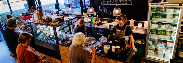 Photo of a cafe with staff behind the counter smiling at their customers. The cafe has an industrial feel to it with black walls, concrete floors and wooden panel on the front of the counter.