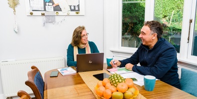 A man writing in a notebook and a woman using a laptop, sitting at a dining room table smiling at each other.