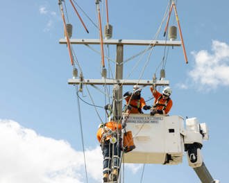 Photo shows two workers in a cherry picker basket with another worker on a ladder with safety harness at the top of a power pole working.