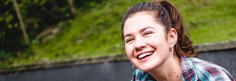 Photo of a young woman outside, crouching down, looking up at something behind the camera, smiling. 