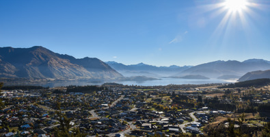 Photo shows Wanaka in the foreground looking toward the lake with the mountains in the background. The sun is shing in the top right corner with a cloudless sky