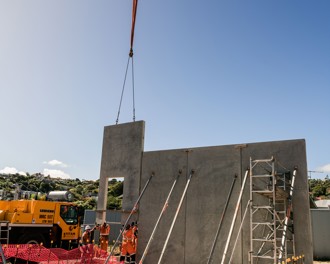 Three sections of a prefabricated concrete wall have been put in place and the crane is lowering a fourth that has a doorway shape in it.  There are steel poles leaning on the wall panels to ensure they stay upright.