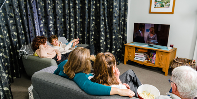 Photo of a family of five sitting on two couches in a living room, watching TV. There is a bowl of popcorn between one of the teenaged girls and grandfather.