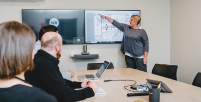 In a conference room with a woman at the front pointing to a large screen that has plans on it.