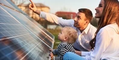 Man shows his family the solar panels on the plot near the house during a warm day.