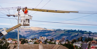 A person in a cherry picker at the top of a power pole working, with city and rural-scape behind them.