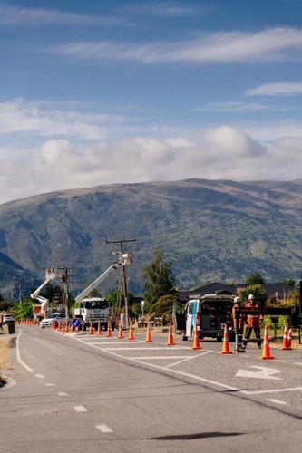Photo shows Delta staff in PPE gear working on power lines and poles with Central Otago mountain in the background