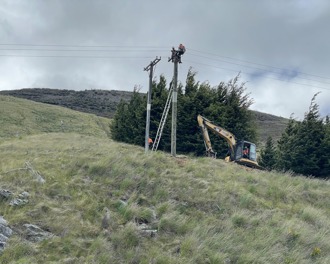 Photo shows two workers at the top of a power pole in the middle of a field on a hillside. There is a CAT digger next to the power pole.