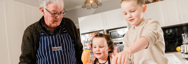 Photo of a grandfather wearing an apron, standing with two young children in the kitchen. They are baking