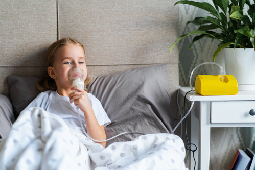 Image shows a small child sitting up in bed holding an oxygen mask over their face. The mask is attached to a yellow box on the white bedside table, alongside a plant.