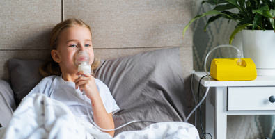 Photo of a young girl sitting up in a grey bed, she is holding an oxygen mask to her face.